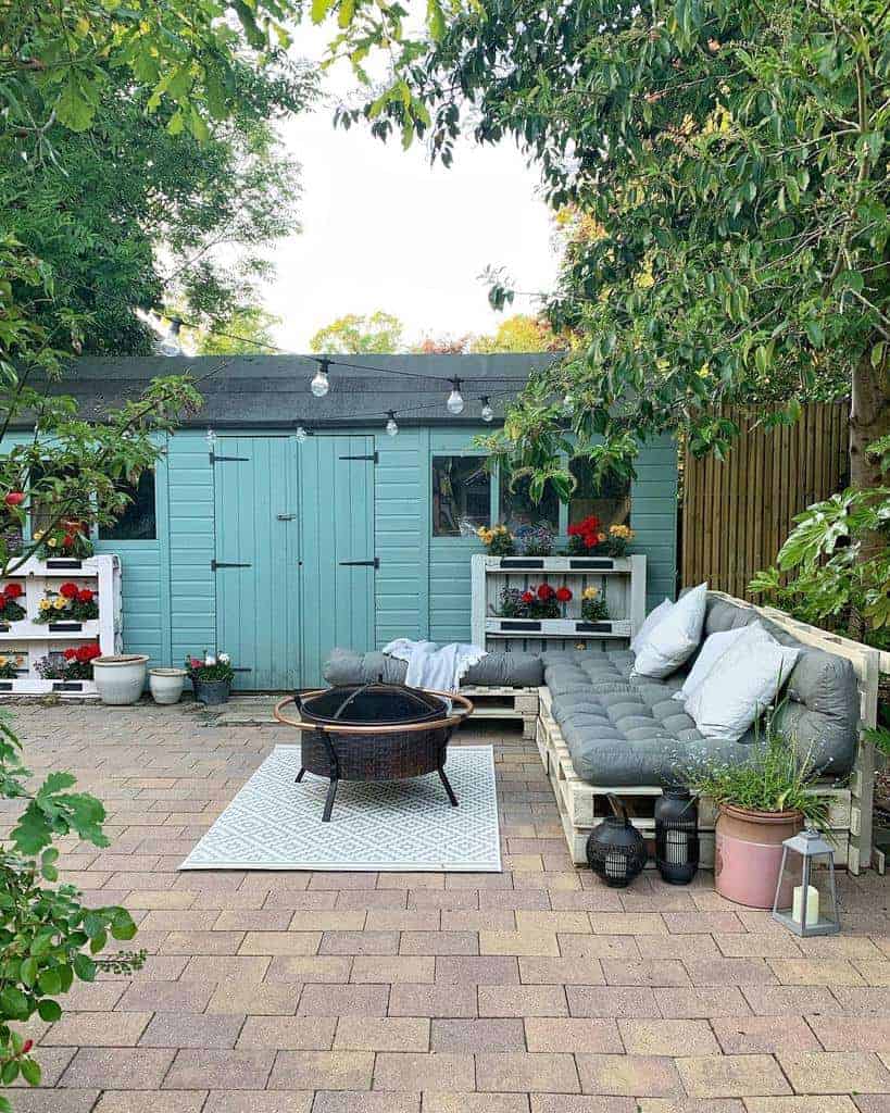 Cozy outdoor pallet sofa with gray cushions, a fire pit, lanterns, and a pastel blue shed surrounded by lush greenery and flowers