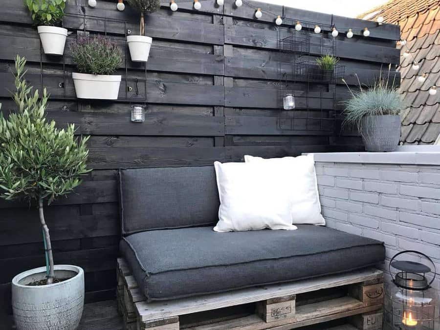 Modern outdoor pallet sofa with dark gray cushions, a black wooden fence, hanging plants, string lights, and a cozy lantern