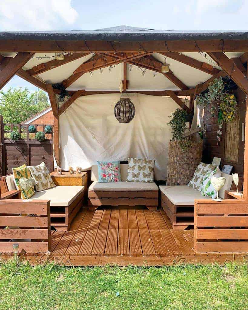 Rustic outdoor pallet seating under a wooden gazebo with beige cushions, decorative pillows, hanging lights, and lush greenery