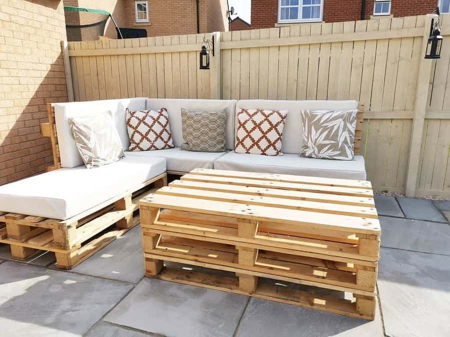 Outdoor seating area with a white cushion L-shaped sofa and patterned throw pillows, alongside a pallet wood coffee table on a patio