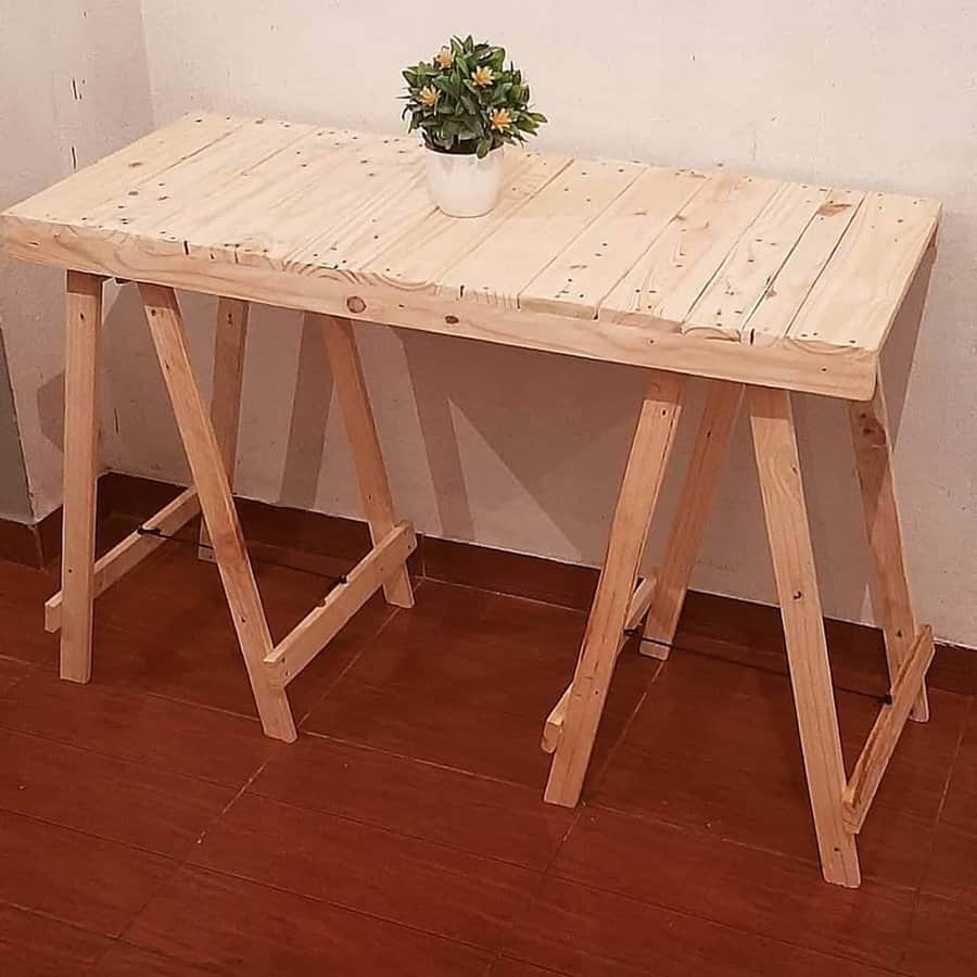 A simple wooden pallet table with a potted plant on top, set against a white wall and placed on a wooden floor