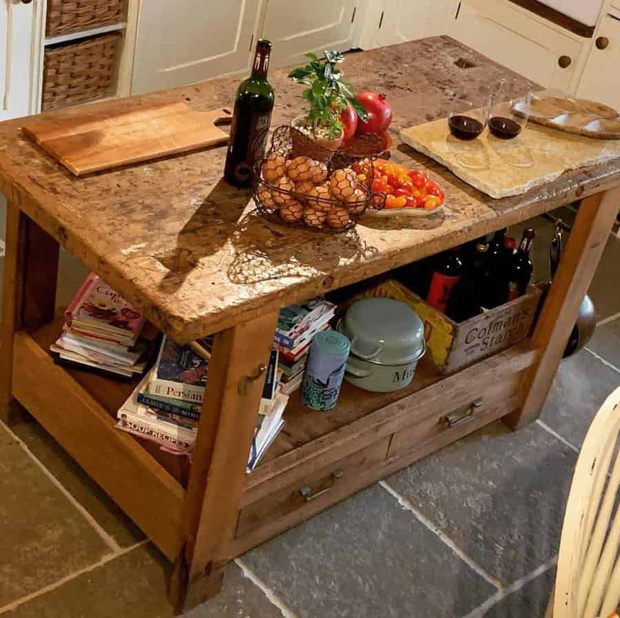 Rustic kitchen island with stone top, holding wine, a basket of eggs, tomatoes, and a magazine shelf underneath, two glasses of wine on top