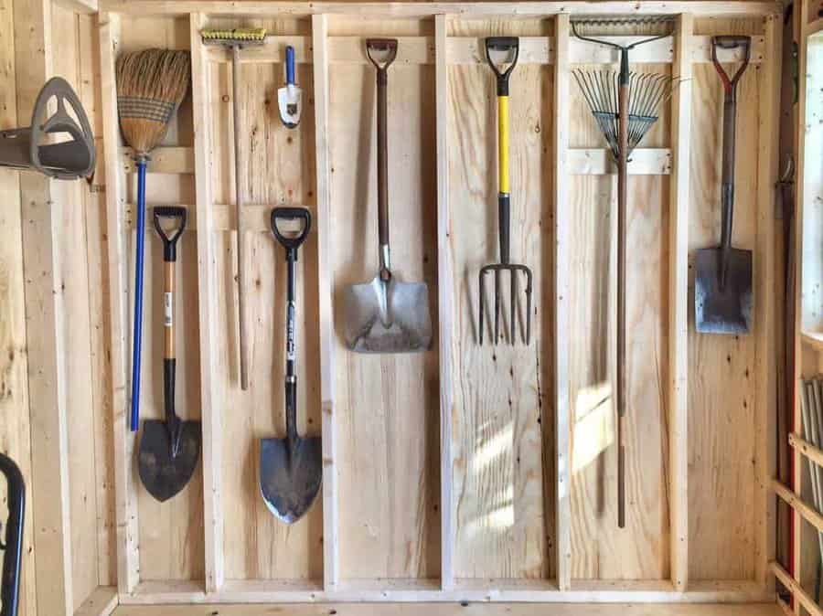 Gardening tools neatly hung on a wooden shed wall, including shovels, rakes, and a broom