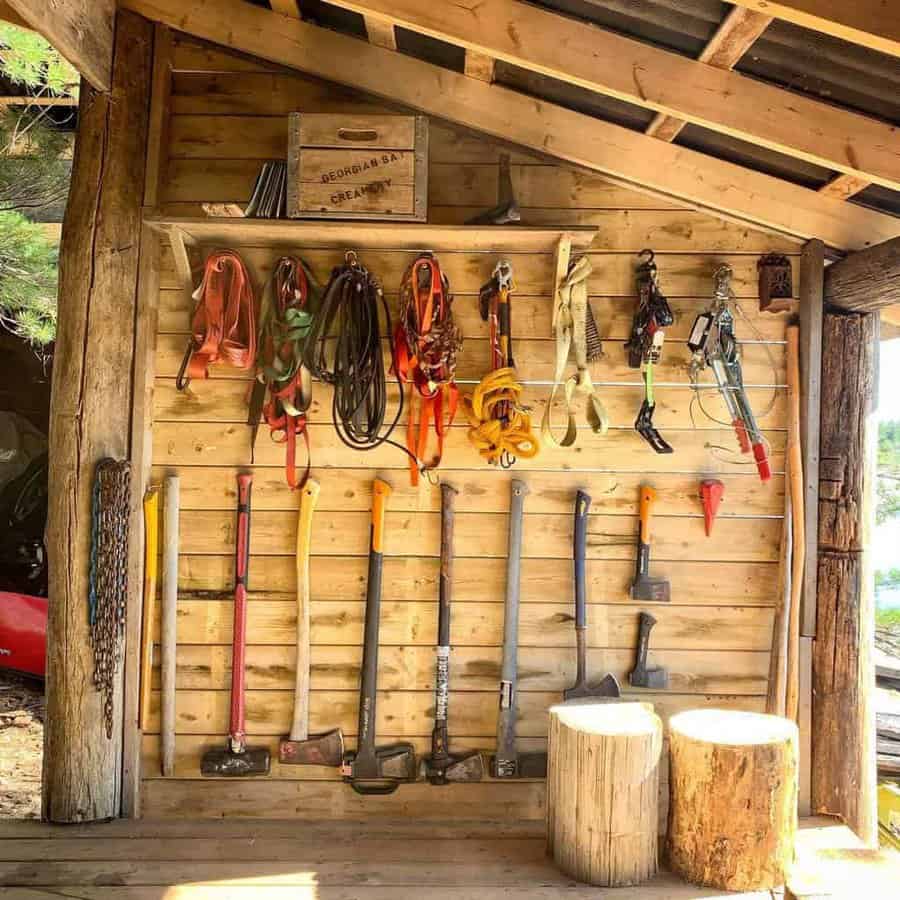 Wooden shed wall with hanging tools: axes, hammers, ropes, and hooks; two tree stump seats in the foreground