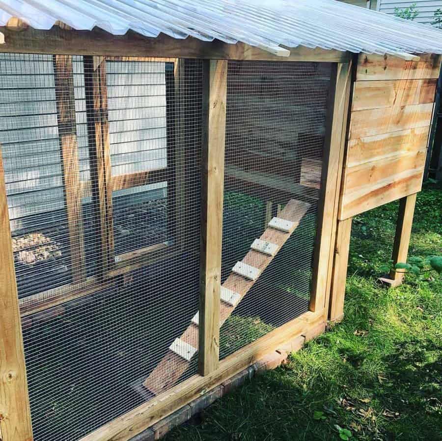 Wooden chicken coop with a slanted roof and wire mesh walls, featuring a small ramp leading inside, situated on green grass