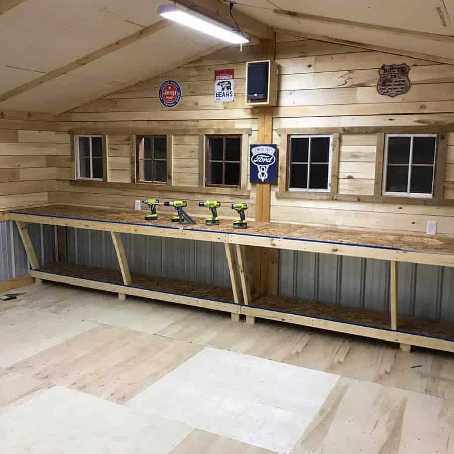 A wooden workshop with a long workbench, power tools lined up, four windows, and vintage signs on the walls