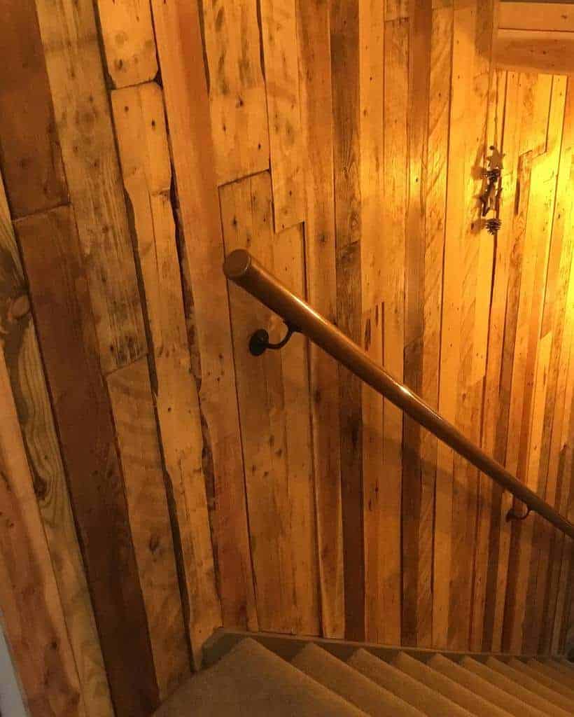 Wood-paneled staircase with a simple handrail, lit by warm, dim lighting; the walls are made of rustic wooden planks