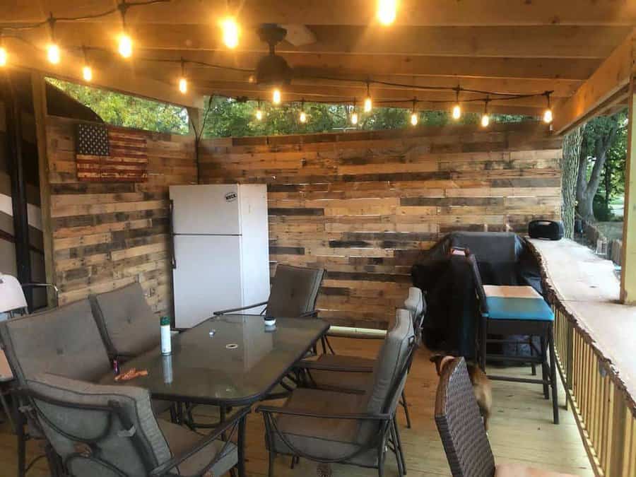 Covered patio with string lights, wooden pallet walls, an American flag, a refrigerator, glass table, chairs, and a railing overlooking trees