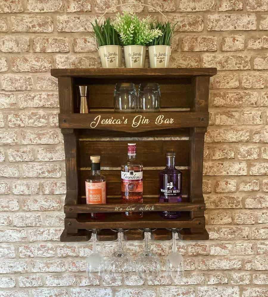 Wall-mounted pallet wooden bar with three gin bottles, glasses, and potted plants labeled "Jessica's Gin Bar" on a brick wall