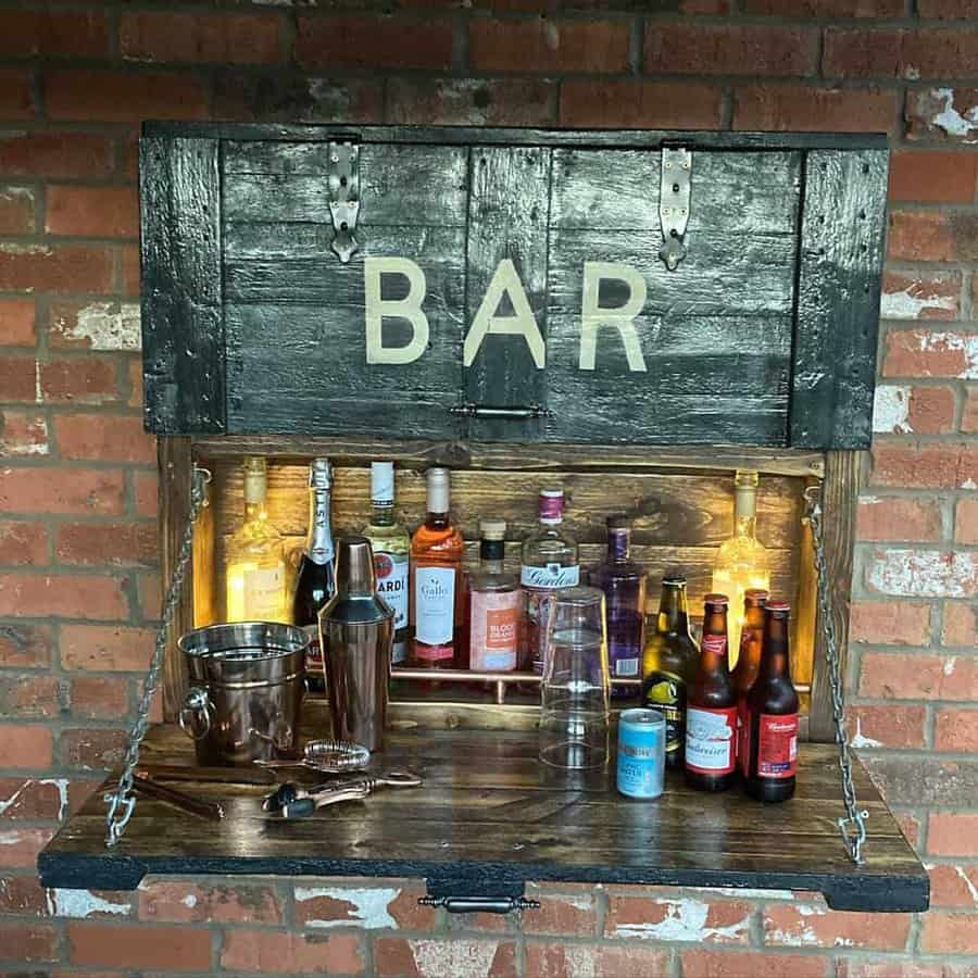 Outdoor wall bar with shelf open, displaying various liquor bottles, beer, and bar tools against a brick wall