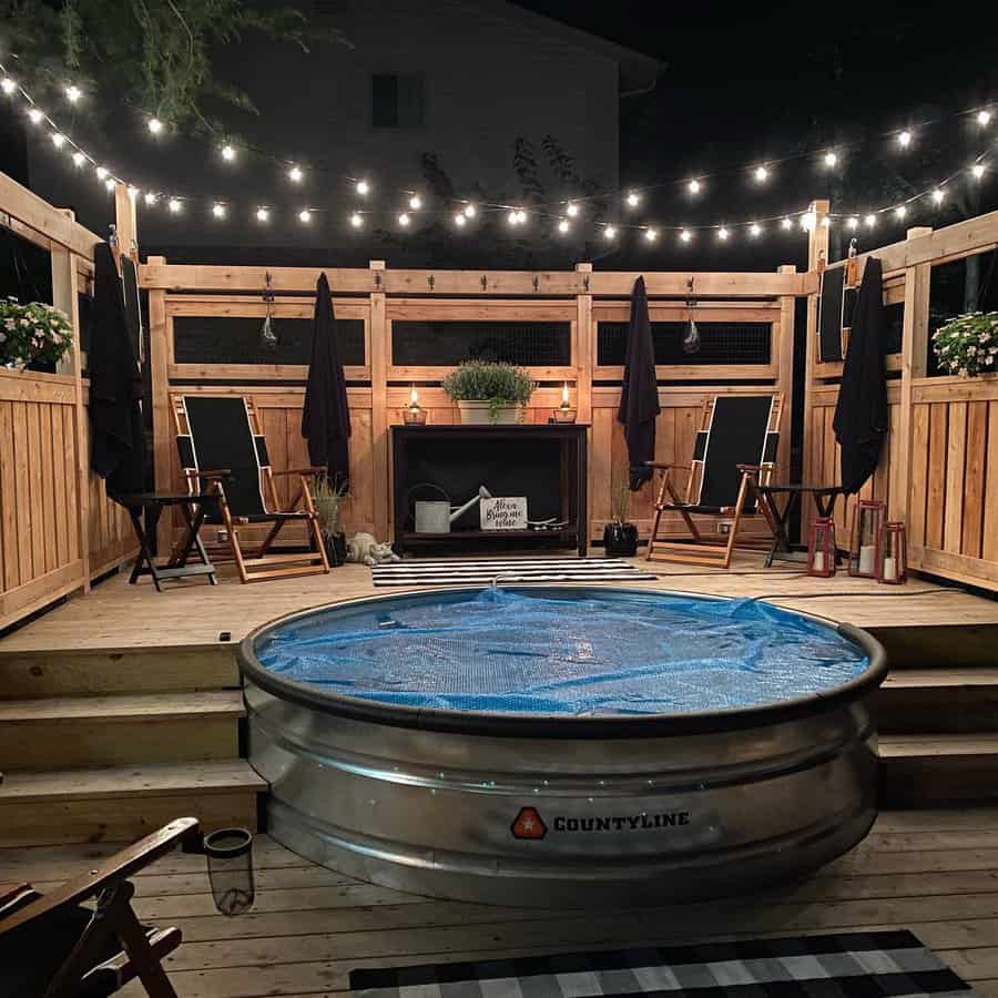 Cozy backyard deck with a stock tank pool, warm string lights, wooden privacy fence, and lounge chairs for a relaxing night retreat