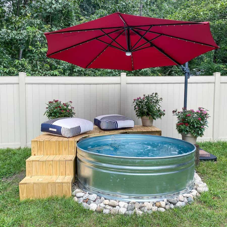 Stock tank pool with wooden steps, cozy cushions, a red patio umbrella, and potted flowers, creating a stylish backyard oasis