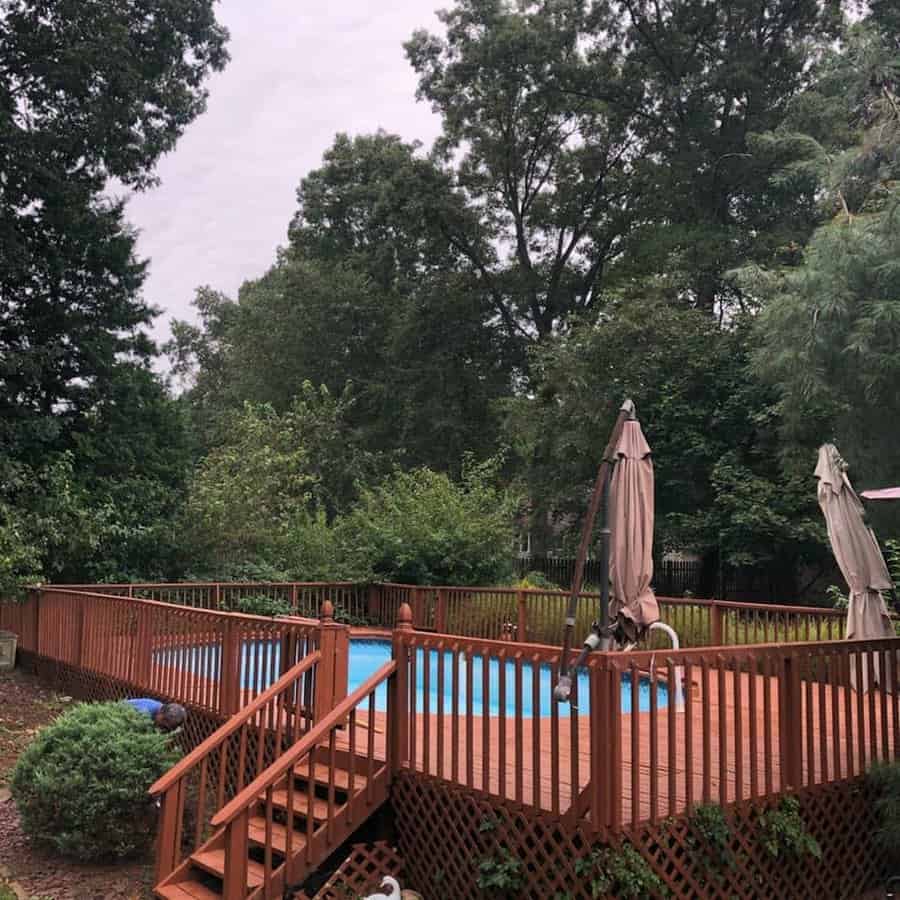 A wooden deck surrounds a swimming pool, with closed umbrellas and lush green trees in the background, and a cloudy sky above