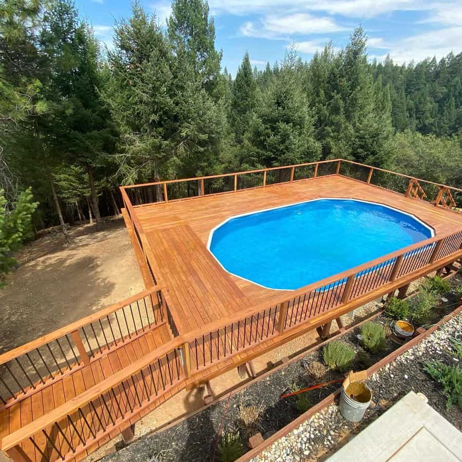 Elevated wooden deck with an oval swimming pool, surrounded by a forest of tall evergreen trees on a sunny day