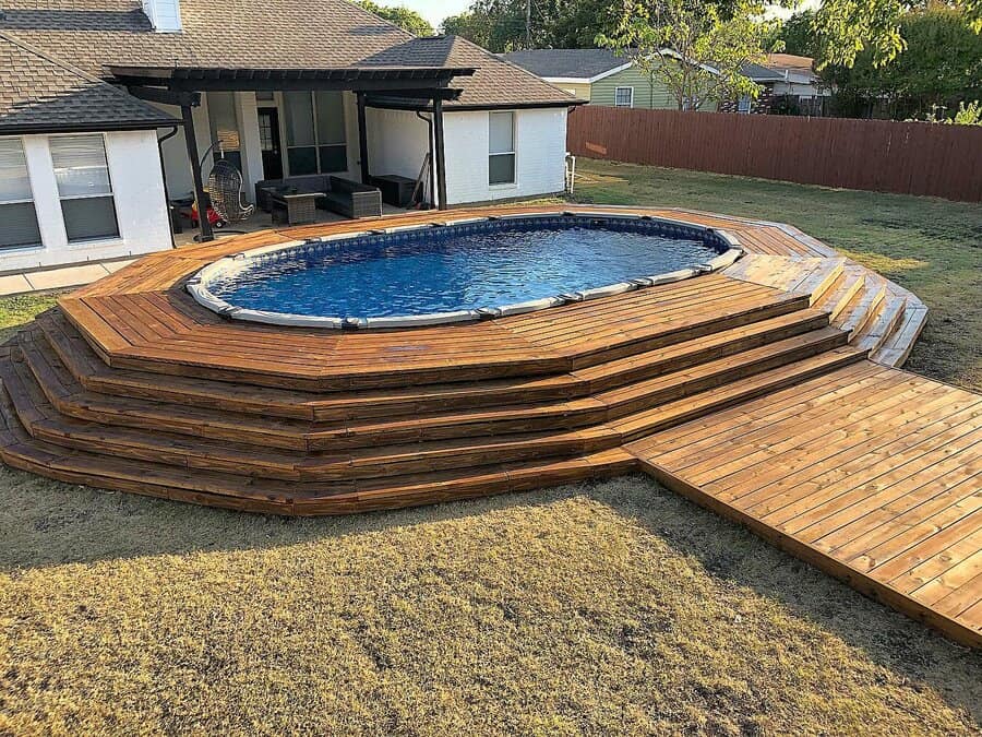 Above-ground pool with multi-layered wooden decking in a backyard next to a white house with a patio