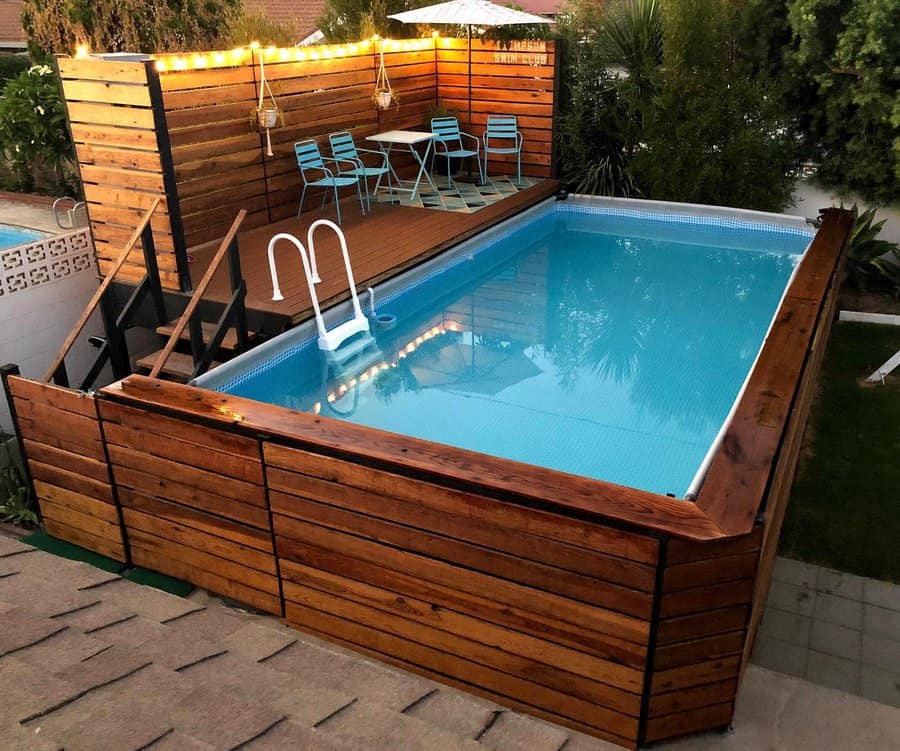 Elevated wooden deck with pool, three blue chairs, a small table, hanging lights, and plants in the background