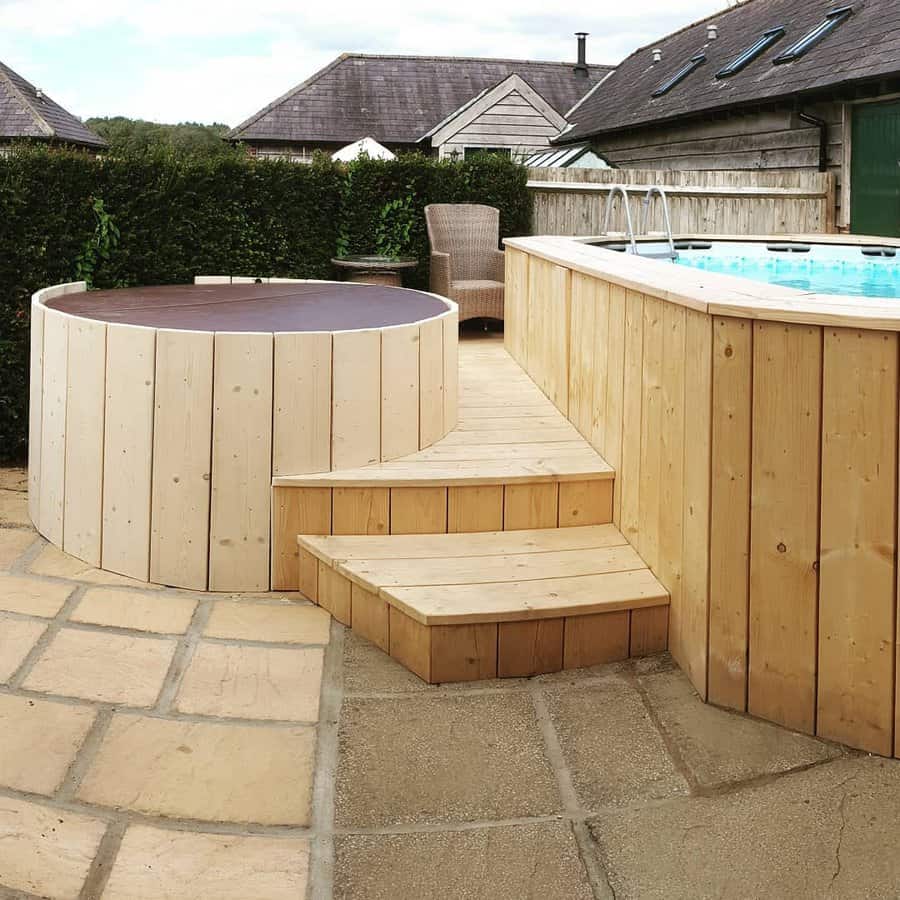 Wooden hot tub and pool with steps on a stone patio, surrounded by greenery and buildings in the background