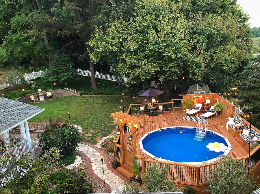 A backyard with a round above-ground pool on a wooden deck, surrounded by trees and a green lawn with a pathway and seating area