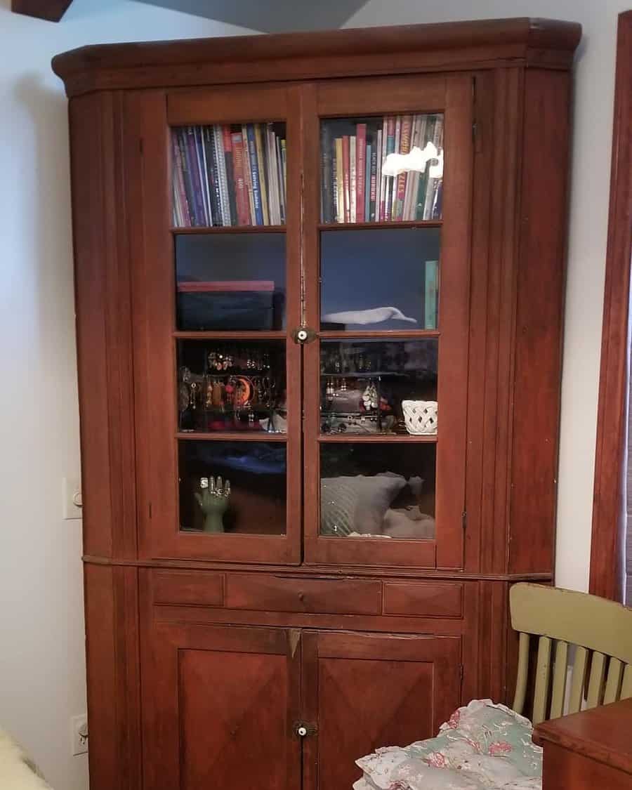 Antique wooden corner cabinet with glass doors, showcasing books, collectibles, and decorative items in a cozy vintage setting.