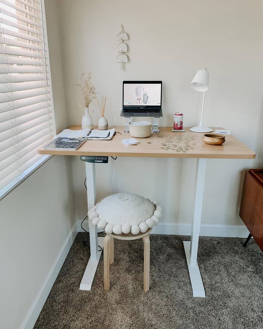 Minimalist home office with a standing desk, laptop, lamp, notebook, and decor; a stool with a cushion is beneath the desk