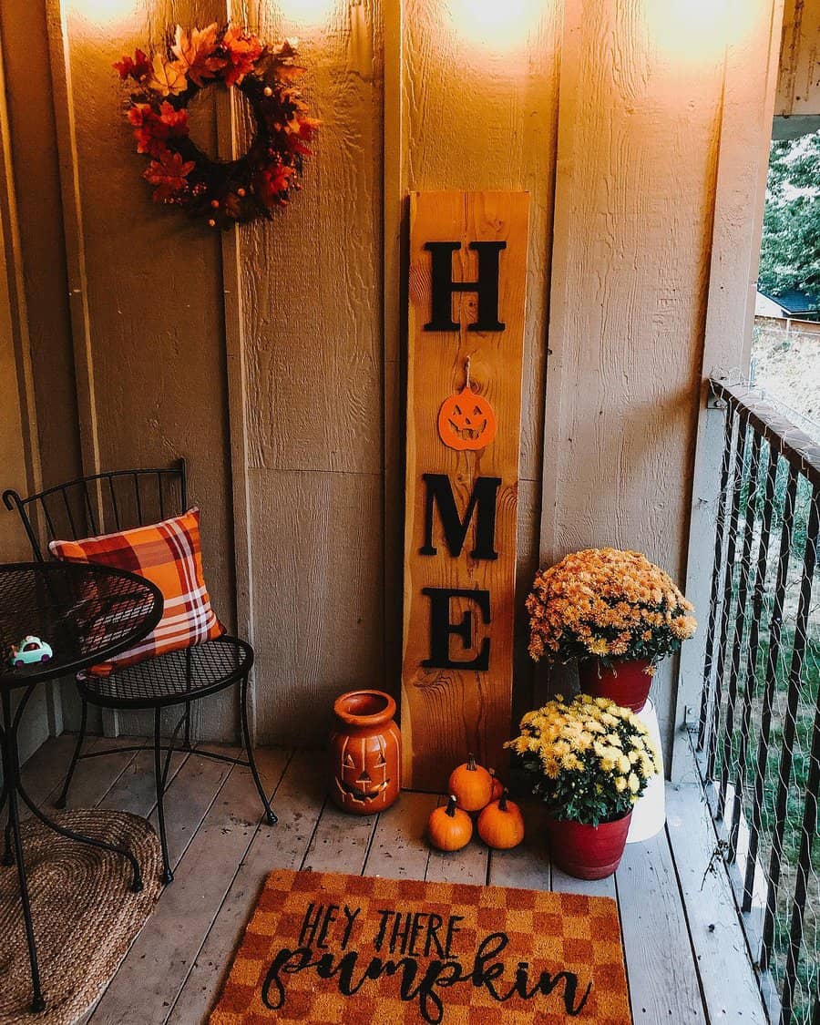 Cozy fall-themed balcony with pumpkins, mums, a 'HOME' sign, a plaid pillow, a wreath, and warm lighting for autumn vibes