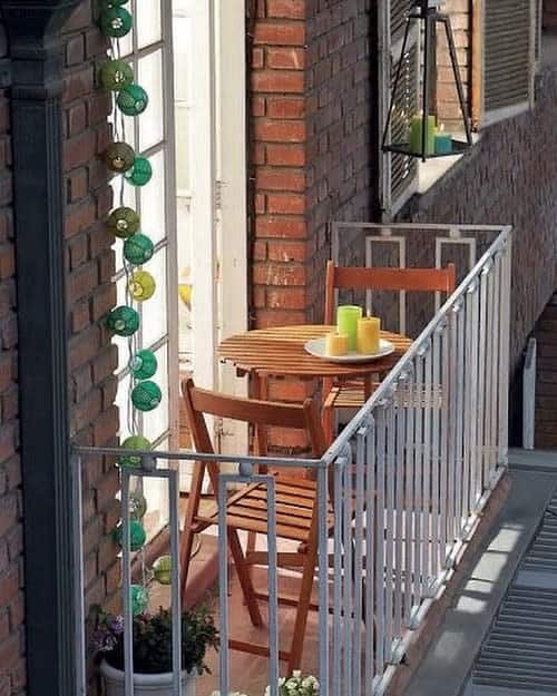 Charming small balcony with a wooden bistro set, green and yellow candles, brick walls, and string lights for a cozy outdoor retreat