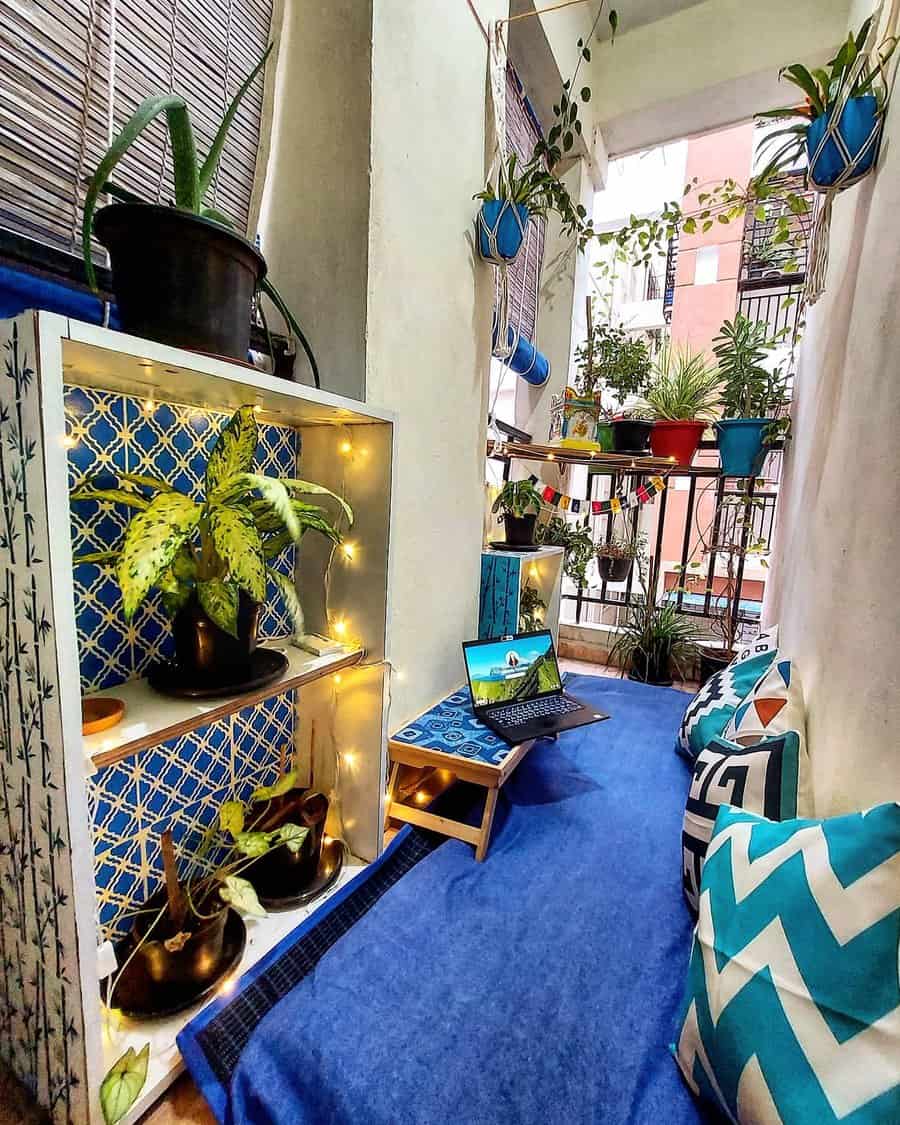 Boho-style small balcony with cozy floor seating, blue decor, patterned pillows, potted plants, fairy lights, and a laptop workspace