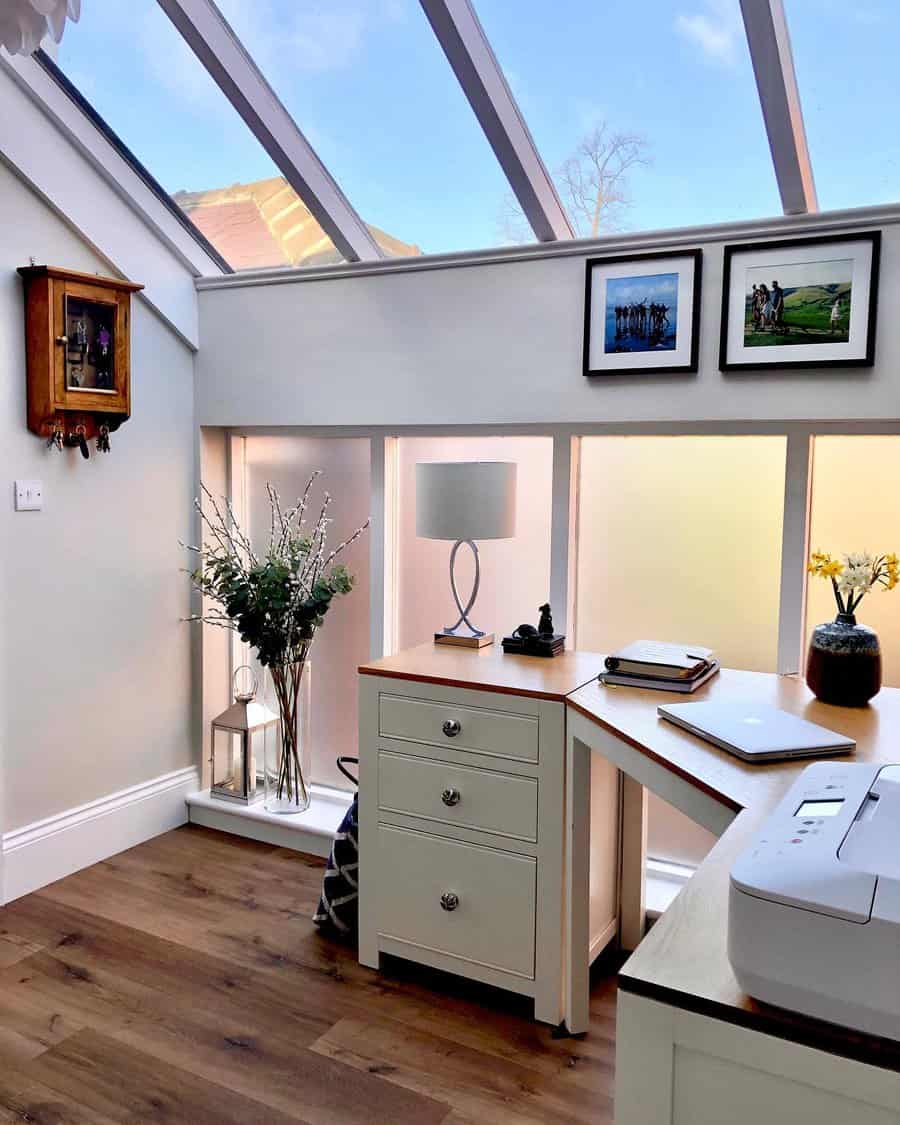 Bright home office with wooden corner desk, skylight, framed photos, and decorative vases. Includes a printer, lamp, and a view outside