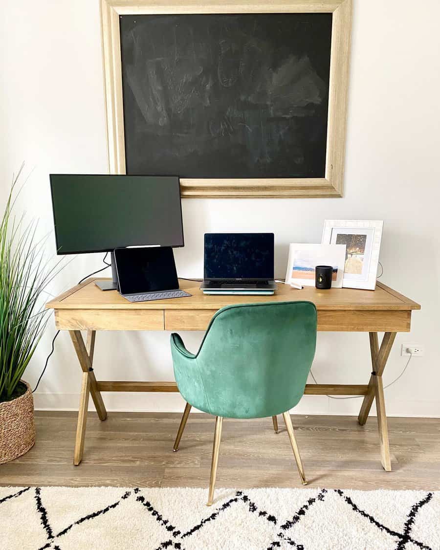 Home office setup with a wooden desk, green velvet chair, dual monitors, a laptop, plant, and a blank chalkboard on the wall