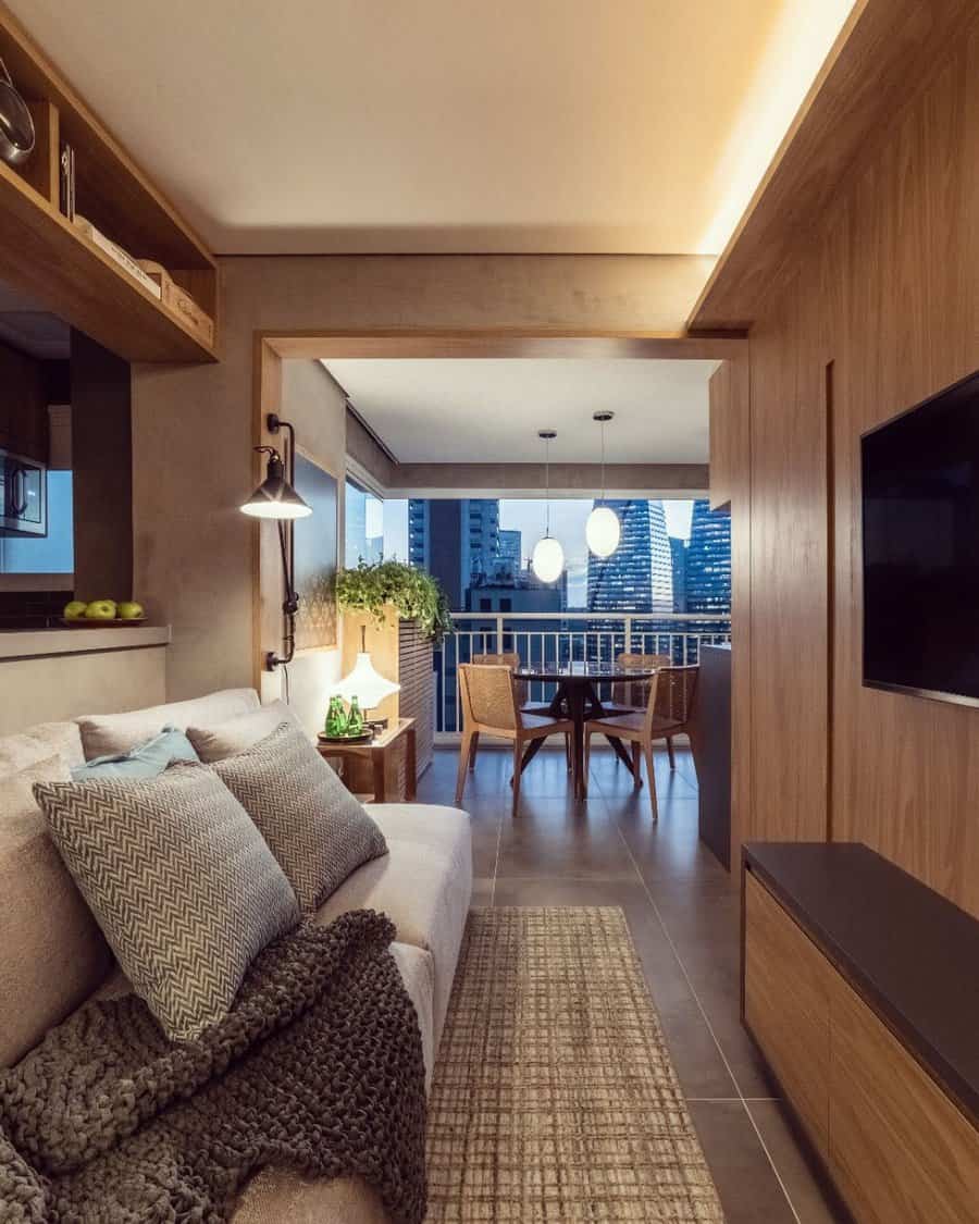 Modern small living room with warm wood tones, a cozy beige sofa, textured pillows, and an open balcony view of the city skyline