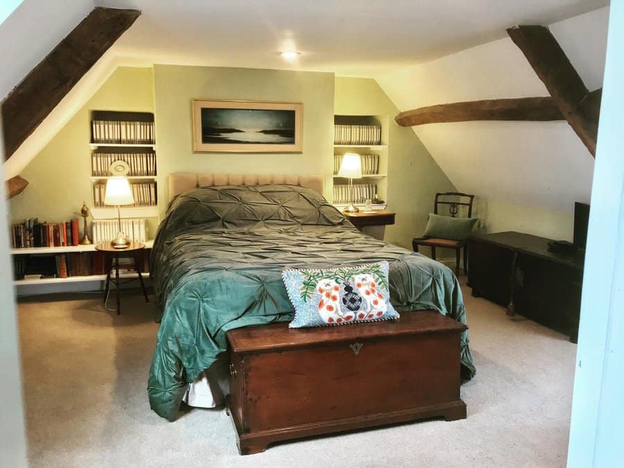 Rustic attic bedroom with exposed beams, built-in bookshelves, and cozy green bedding. A vintage trunk adds charm and storage.