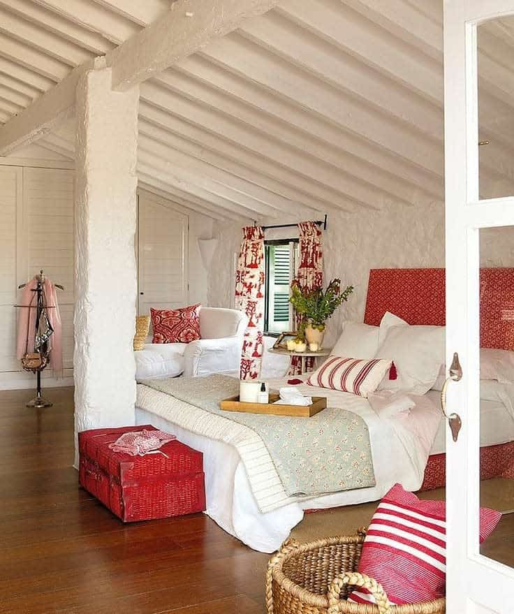 Bright attic bedroom with whitewashed beams, red accents, and cozy layered bedding. Rustic charm meets warmth with soft textiles and wood tones.