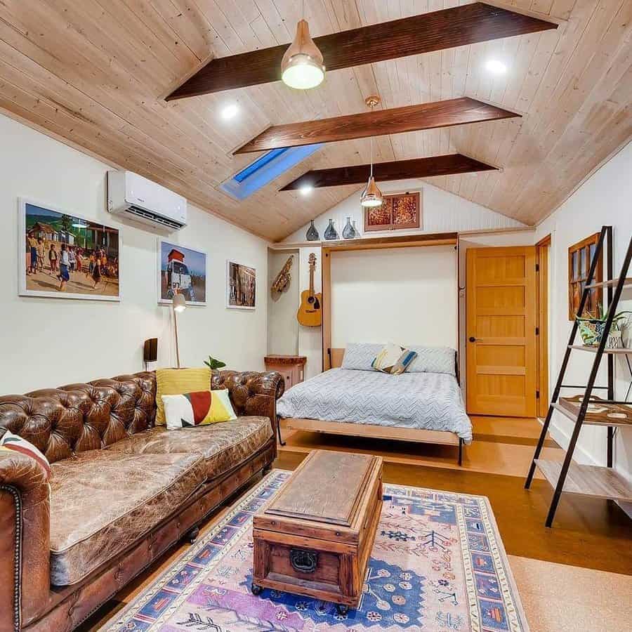 Rustic attic bedroom with exposed beams, a leather Chesterfield sofa, and a Murphy bed. Warm wood tones and eclectic decor create a cozy vibe.