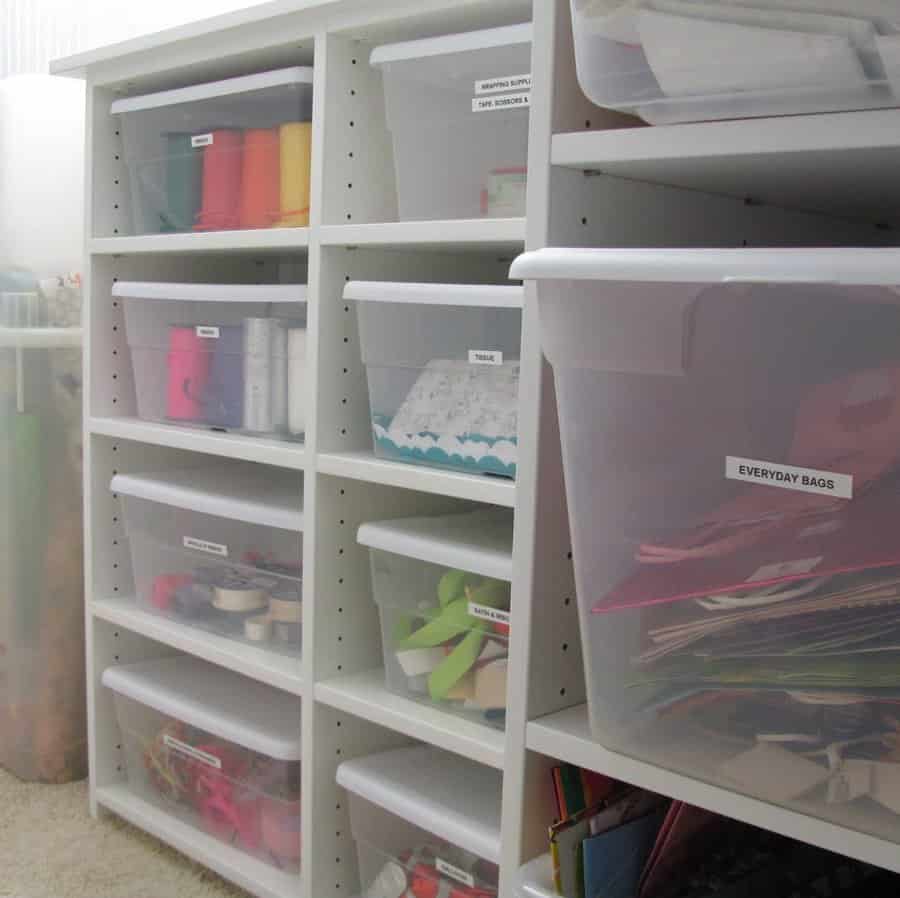 Clear plastic bins with labeled craft supplies neatly stored on white shelves in an attic, creating an organized and clutter-free space.