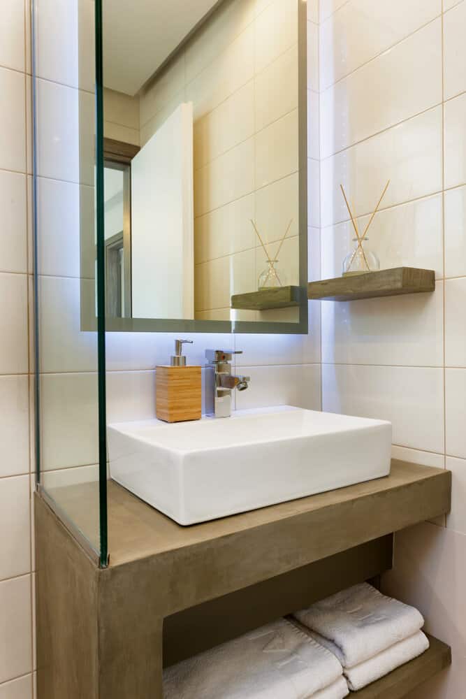 Modern bathroom sink with a large mirror, soap dispenser, and reed diffusers on shelves; towels are stacked underneath