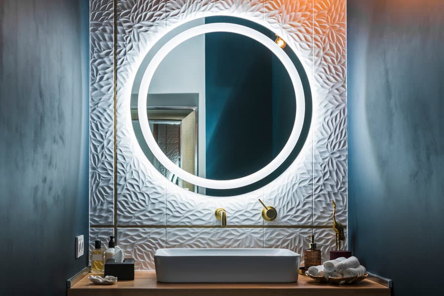 Modern bathroom with dark walls, a circular illuminated mirror, textured white tile backsplash, and a sleek sink with gold fixtures