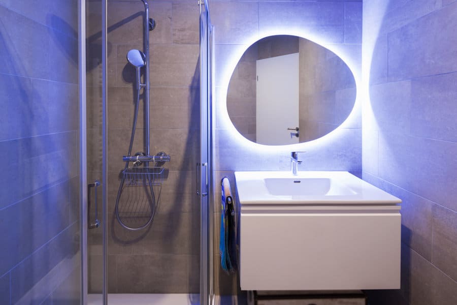 Modern bathroom with glass shower and illuminated oval mirror above a white sink vanity, gray tiled walls and a closed white door