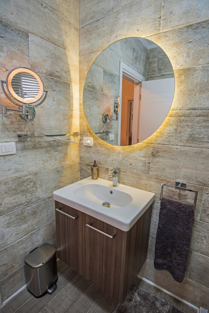 Modern bathroom with a round-lit mirror, wooden vanity, white sink, and towel on a holder featuring a wall-mounted mirror with magnification