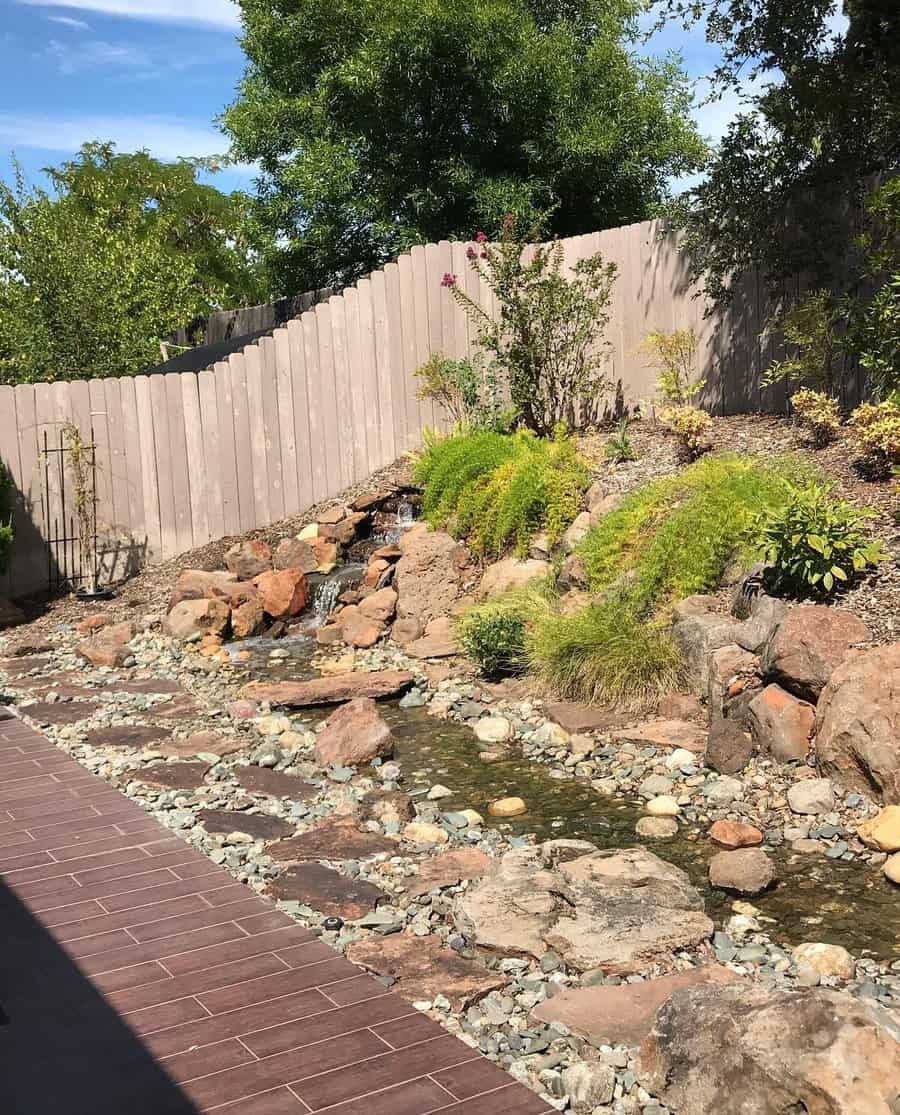 Rocky garden with a small stream, surrounded by greenery, and a wooden fence in the background