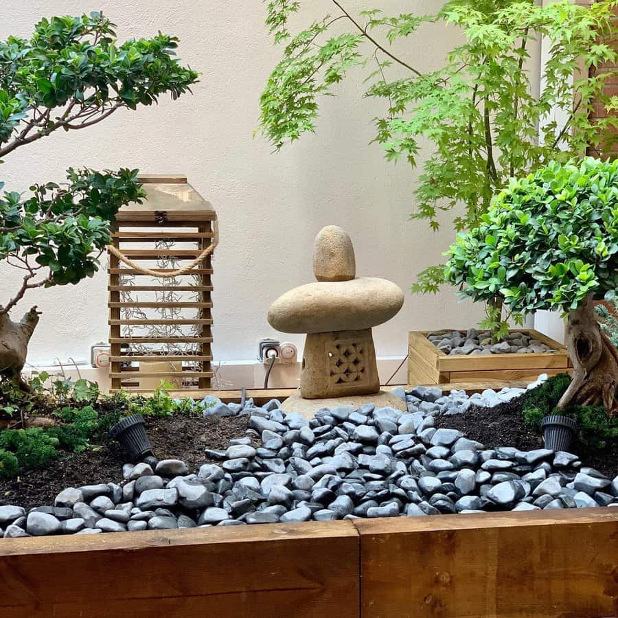 A small stone lantern is surrounded by greenery, potted trees, and gray pebbles in a wooden planter against a white wall