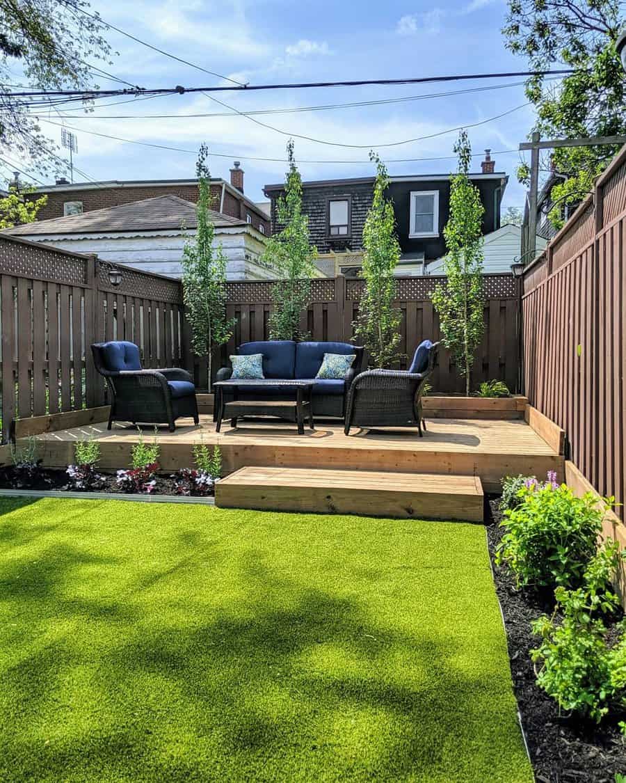 Cozy backyard with wooden deck, wicker furniture, green lawn, flowerbeds, and tall plants against a wooden fence