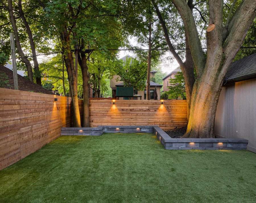 Modern low-maintenance backyard with artificial turf, wooden privacy fence, accent lighting, and large mature trees