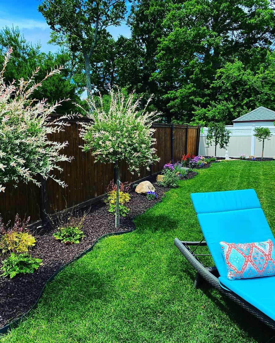 Vibrant backyard with mulched garden beds, decorative trees, lush lawn, and a bright blue lounge chair with a patterned pillow