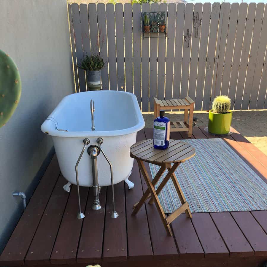 Outdoor bathtub on a wooden deck with a folding table, striped rug, and potted cacti against a fence backdrop