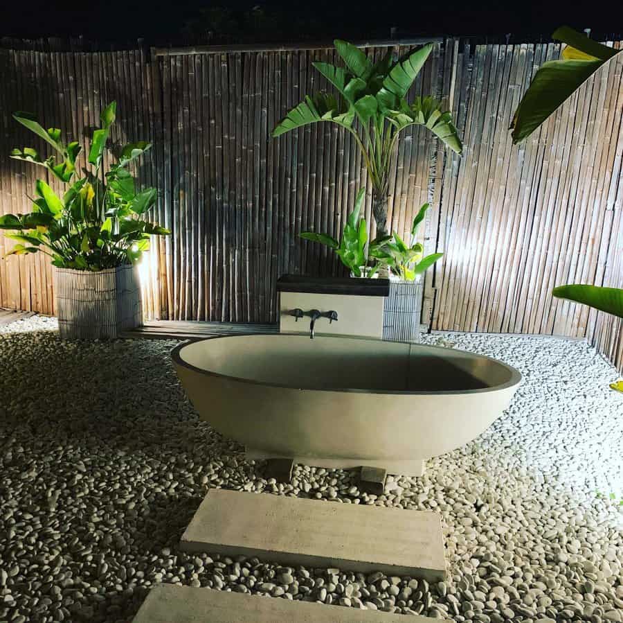 Outdoor stone bathtub on pebble ground, surrounded by lush green plants and bamboo fence, illuminated at night