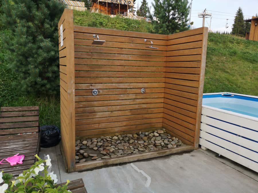 Outdoor wooden shower stall with stone floor, next to a white container pool, surrounded by greenery and a wooden bench