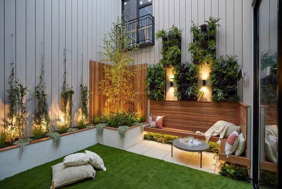 Cozy patio with greenery, wooden seating, cushions, and a small round table on a tiled floor and grass area; vertical garden wall with lights