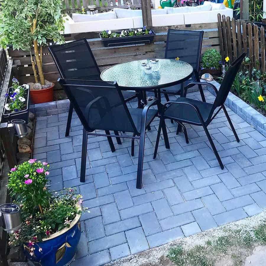 A garden patio with a round glass table and four black mesh chairs on a gray brick surface, surrounded by plants and wooden fencing