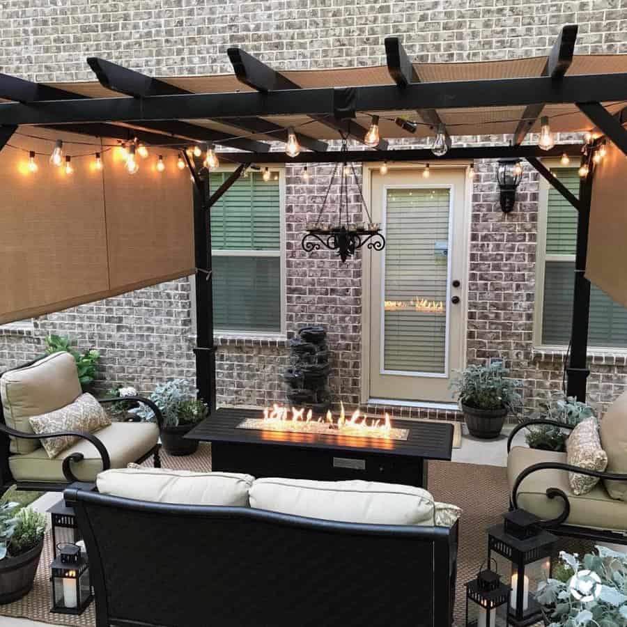 Cozy patio with a pergola, string lights, seating, and a fire table; brick wall backdrop and potted plants enhance the setting