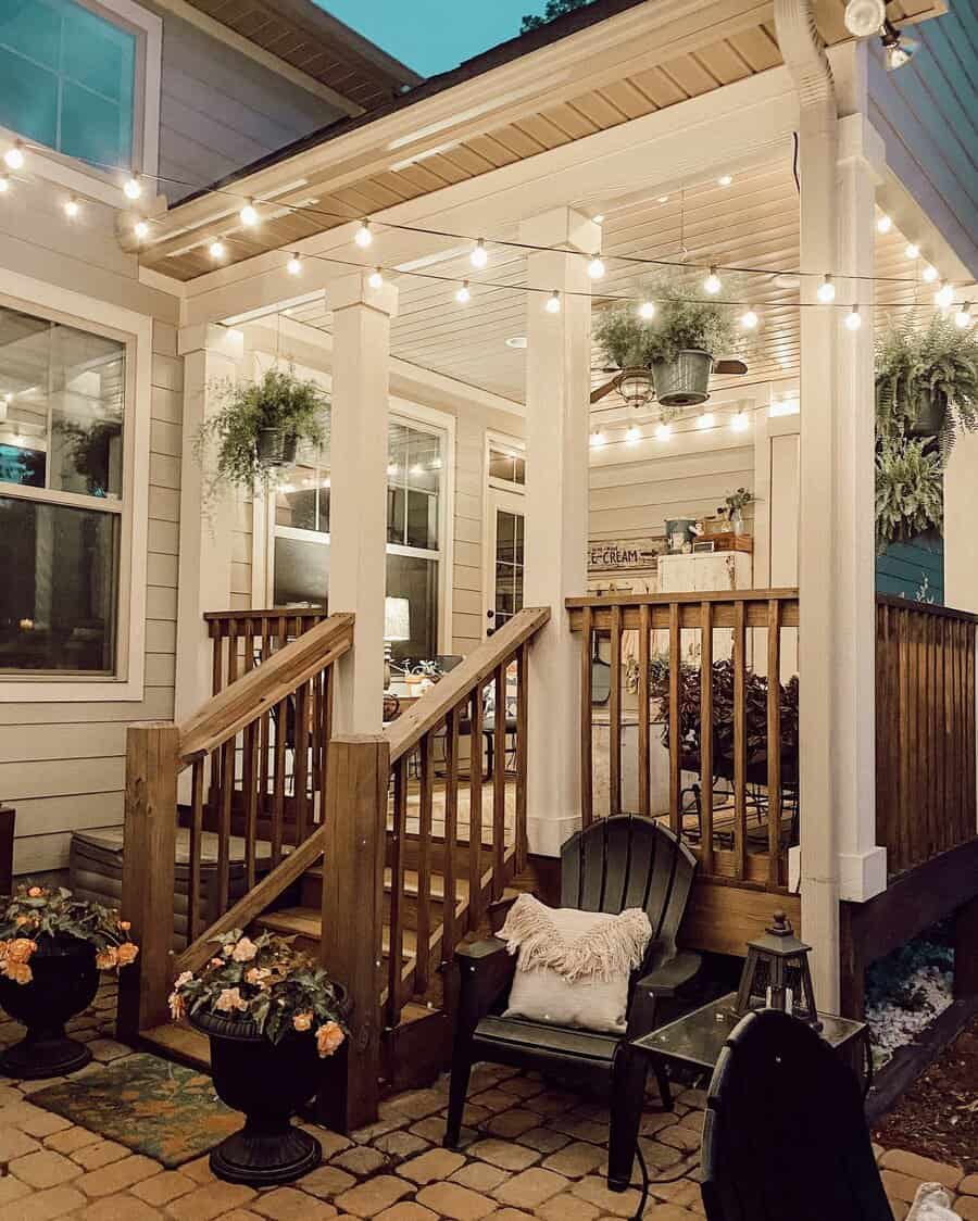 Cozy porch with string lights, potted flowers, ferns, and wooden furniture, including chairs and a table, against a house backdrop